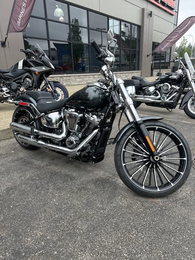 This image captures a close-up view of a black Harley-Davidson motorcycle parked on an asphalt surface outside a building. The motorcycle is the primary subject, showcasing its chrome details, black paint, and a large front wheel with a distinctive black and silver spoke design. A second, partially visible motorcycle is parked behind it.
The setting appears to be a dealership, indicated by the large glass windows of the building and a banner in the background that reads "BARNES HARLEY-DAVIDSON". The building has a modern facade with gray siding and stone accents. The time of day is difficult to determine precisely, but the lighting suggests it could be daytime, possibly overcast given the somewhat muted reflections in the windows. No people are visible in the image. The overall mood is still and commercial, focusing on the display of the motorcycles.