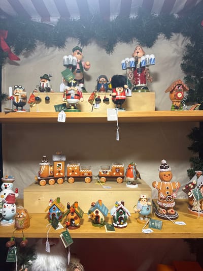 A shelf displays Christmas ornaments. On the left, are German nutcrackers. On the right, a gingerbread train, decorative birdhouses, and snowmen. The shelf is wooden and partially framed by a green garland. Tags hang from the ornaments, indicating their prices. Some items depict people, like the gingerbread man, nutcracker soldiers, and a nurse. Other items are nature-themed, like the birdhouses, cat, and snowmen.