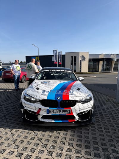 A white BMW M4 or similar model "Safety Car" with extensive racing livery is parked prominently on a light grey interlocking paver surface, facing the viewer. The car features BMW Motorsport stripes (blue, dark blue, red) extending from the grille over the hood, and is covered with repeating "BIMMER TUNING" and "official car of BIMMER TUNING" text. "Safety Car" is emblazoned in red on the hood. The vehicle's front displays a German license plate "BM WP 9991" and aggressive aftermarket aerodynamic elements including a large front splitter and canards.
In the mid-ground, to the left of the car, three adult males are standing and conversing. One man with glasses wears a light t-shirt, another a light t-shirt with a green "VULCAN ALOHA" graphic, and the third a white t-shirt. Behind them, a red sedan, possibly a Tesla, is partially visible.
The background features modern commercial buildings under a clear, bright blue sky. A distinct light-colored building displays vertical dark window elements and a large, dark eagle or bird of prey logo on its facade. Behind this, a darker-colored building with large windows is visible. Two tall dark banners, one clearly reading "PAUL CARS GERMANY," are positioned between the people and the buildings. The scene is illuminated by direct sunlight, casting shadows. This location is identified as within the Nürburgring complex in Meuspath, Germany, specifically near the Ring°Boulevard or Ring°Werk area.