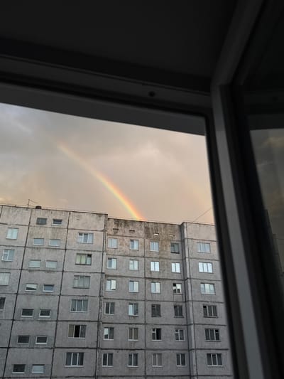 The image shows a view from a window looking out at a gray, multi-story apartment building and a sky with a faint double rainbow. The building is made of concrete panels and has numerous rectangular windows, some of which appear to be reflecting the sky. The sky is overcast with a yellowish hue, suggesting it is either sunrise or sunset. The framing of the window in the foreground, which is dark and appears to be a window frame, suggests the photo was taken from inside a room. The architecture of the building, characterized by its utilitarian design and visible concrete panels, is typical of Soviet-era mass housing prevalent in cities like Norilsk, Russia. The presence of a rainbow indicates recent rainfall or atmospheric moisture. No people are visible in the frame, and the overall scene depicts a somewhat stark urban environment under a dramatic sky.