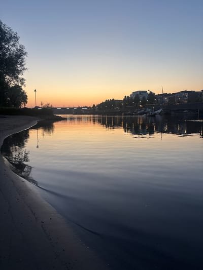 This is a photograph of the Rhine river at sunset in Arnhem, Netherlands. The sky is a gradient of light blue to orange, reflecting on the calm water. On the left bank, a sandy shore is visible with a single tree and a lamppost. Across the river, the Arnhem skyline is dotted with buildings and trees, with a bridge spanning the water in the distance. Several boats are docked along the riverfront. The overall atmosphere is serene and peaceful.