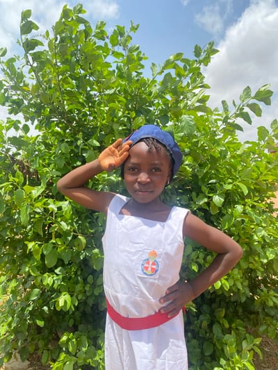 A young girl stands in front of a lush green bush under a partly cloudy sky in Turu, Nigeria. She wears a blue hat, a white uniform with a "The Girls' Brigade" logo on the chest, and a red sash around her waist. She is posing for the camera, smiling with her left hand raised.