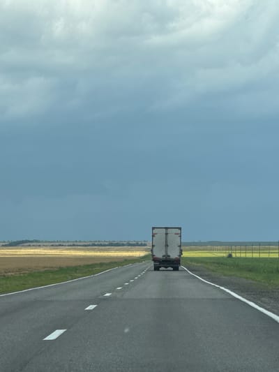 A white box truck is traveling away from the viewer on a two-lane asphalt road under a cloudy, overcast sky. The road has a white solid line on the right edge and dashed white lines marking the lanes. The landscape to the left and right of the road is agricultural, with fields of golden crops and green grass. In the distance, a treeline borders the fields. The truck is centered on the road and appears to be moving forward. The sky is a muted blue with darker clouds forming. There are no people visible in the image. The scene is one of rural travel on a highway.