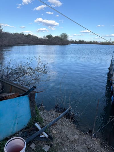 The image presents a view of a body of water, possibly a lake or river, in or near Rozovo, Bulgaria. The sky is blue with scattered clouds. A line stretches across the frame, possibly a cable or rope. The shoreline is visible on the left with vegetation and trees. The water reflects the sky. On the right, the shoreline is closer and covered in dirt, grass, and debris, including pipes and a blue structure that appears to be abandoned. The scene is disorganized, suggesting an area where there has been dumping or neglect. No people are visible.