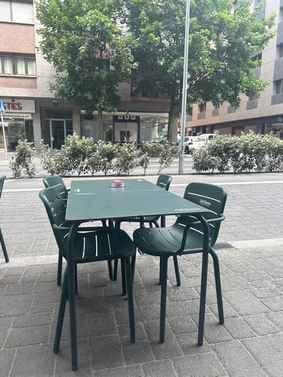 The image displays an outdoor cafe seating area located on a paved sidewalk alongside an urban street. The perspective is slightly elevated, looking across and along the sidewalk.
In the foreground, a dark green plastic table is surrounded by matching green plastic chairs. Some chairs are stacked, suggesting the establishment might be closed or preparing. A white and red ashtray rests on the table. The sidewalk features grey paving stones. To the left, a street with vehicles, including a partially visible white car, runs parallel to the sidewalk. A line of well-maintained green bushes separates the seating area from the road.
Beyond the street, multi-story buildings with modern architecture, some with checkered grey facades and others with large windows, form the backdrop. Lush green trees are visible along the street, blending with the buildings. The bright, even lighting indicates it is daytime, likely sunny. No people are visible, and no specific activity is taking place beyond the arrangement of the cafe furniture.
Visible text provides several clues. On the green chairs and table, the brand "Damm" and "Volt Damm DOBLE MALTA" are clearly legible, referring to a Spanish beer. On one of the buildings across the street, the word "BAR" is seen. Another sign further down displays "ESTES" and an email address "tustes@andorra.ad", confirming the location as Andorra, as specified. A "P" symbol, likely indicating parking, is also visible on a building.
