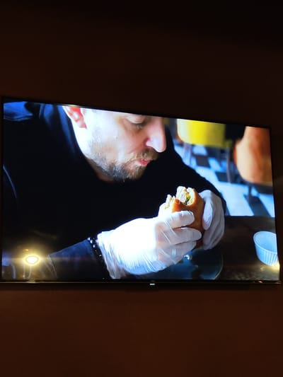 A medium shot captures a man wearing disposable gloves and a black hoodie, holding a partially eaten sandwich. The man has short, light brown hair, a beard, and appears to be in his late 30s or early 40s. He is seated at a table with a dark wooden surface. To his right, a small white paper cup is visible. In the background, a blurred checkered floor and yellow and orange seating can be seen, suggesting an indoor dining environment, possibly a cafe or restaurant. The image is a photograph of a television screen. The man is looking down at the sandwich. The television is mounted on a dark brown wall.