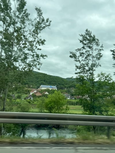 The image, likely taken from a moving vehicle (train), shows a view of the landscape in Raska Municipality, Serbia. The foreground contains parts of a tree and a metal rail. Beyond that, there is a river or stream adjacent to a grassy field. A cluster of buildings sits amongst trees on the slope of a mountain. The sky is overcast.