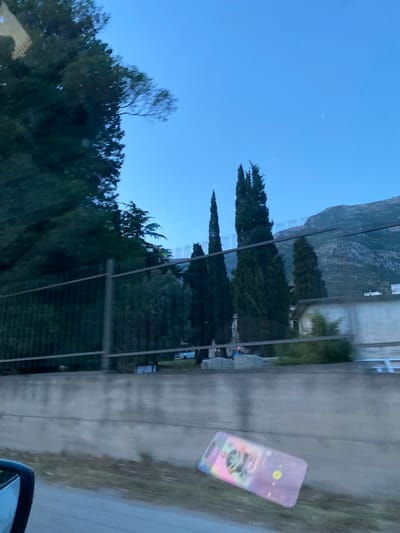 The image shows a view from a vehicle window, likely a car, in what appears to be a rural or semi-rural area in or near Zgrade, Montenegro. The sky is light blue, suggesting early morning or late afternoon. Tall, slender trees resembling Italian cypresses are visible, leading towards a mountainside in the distance. A multi-story building, partially obscured, is visible on the right. In the foreground, on the right side of the window, a smartphone is visible, with its screen displaying music or audio content. The vehicle's side mirror is partially visible in the lower right corner. The road or path adjacent to the trees appears to be made of concrete or asphalt. There are no visible people.