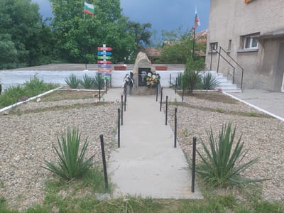 The image shows a memorial site outside a building in the village of Blajievo, Bulgaria. The memorial features a large rock with a black and white portrait of a man, inscribed with "Васил Левски 1837". Wreaths of flowers are placed at the base of the rock. A concrete pathway leads to the memorial, lined with black metal posts and chains. Surrounding the pathway is a gravel-covered area with several spiky green plants and some grass. Behind the memorial, a flagpole displays the Bulgarian flag. In the background, there is a multi-story building with a rough, textured facade and stairs leading up to an entrance. Trees and some houses are visible beyond the building. Several colorful directional signs are placed to the left of the memorial, pointing towards different virtues or qualities, such as "добрина" (kindness) and "успех" (success). The sky is cloudy.