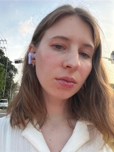 A close-up selfie shows a young Caucasian woman with light brown hair and fair skin, wearing a white collared shirt. She has a purple wireless earbud in her left ear and a small hoop earring in her left earlobe. Her right cheek is flushed, and her lips are slightly parted, revealing a glossy appearance. The background suggests an outdoor setting in Pocheon-si, South Korea, with utility poles and wires visible, along with trees and a white van in the distance. The sky is a pale blue with some clouds.