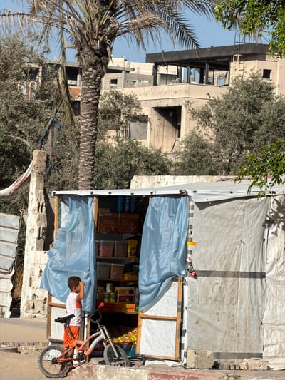 In Jabalia, Palestinian Territories, a young boy stands near a small roadside kiosk. The boy, wearing an orange t-shirt and shorts, is positioned to the left of the kiosk, looking upwards and to his left. A small bicycle, a child's model with orange accents, is placed in front of him, leaning against the kiosk.
The kiosk itself is constructed with a white tarp for a roof and sides, with light blue tarpaulin serving as a curtain or cover for its front opening. Inside, shelves are stocked with various goods, including what appear to be packaged snacks and food items. The kiosk is framed with wooden supports.
In the background, evidence of damage and reconstruction is visible. A palm tree stands to the left of the kiosk, and behind it are buildings with a damaged appearance, suggesting recent conflict or decay. The structures show signs of being incomplete or damaged, with exposed concrete and broken windows. The overall environment appears to be an urban setting, with elements of daily life juxtaposed with signs of destruction.