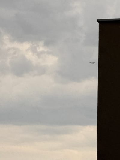 The image shows a cloudy sky with an airplane in flight. A dark building occupies the right side of the frame. The scene is set in the city of Trap, Montenegro, though no specific landmarks are visible to pinpoint the exact location. The lighting suggests it is either early morning or late afternoon, given the subdued light and overcast conditions.