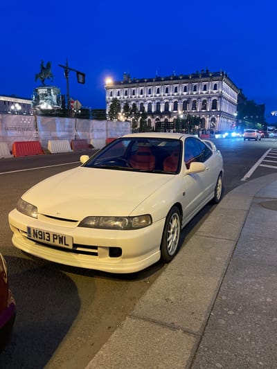 The image depicts a white Honda Integra DC2 coupe parked on a street at dusk in Vaganovo, Russia. The car is positioned in the foreground, facing slightly to the right, with its front bumper and driver's side visible. The vehicle has a British license plate that reads "N913 PWL" and red interior seats.
Behind the car, a row of white and red construction barriers lines the sidewalk. In the middle ground, a large, ornate building with multiple arched windows is illuminated, suggesting it is a historical structure or a place of importance within the city. A street light and a traffic signal are visible near the building. Further in the background, an equestrian statue on a pedestal stands against the twilight sky. Other vehicles, including a white SUV and a dark car, are visible in the distance on the road, indicating traffic activity. The sky is a deep blue, transitioning from the day to night. The overall environment suggests an urban setting, likely a public square or a busy street in Vaganovo. There are no discernible people in the immediate vicinity of the car, though some figures may be present near the illuminated building.
