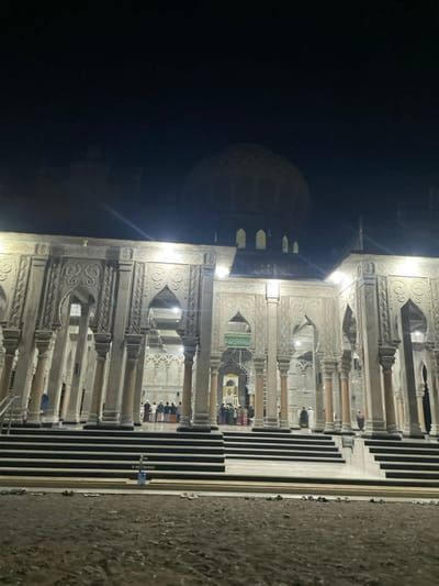 A night shot reveals a mosque-like structure, predominantly white with intricate arched designs. The building features a central dome and is illuminated, with figures appearing to pray inside. Stairs lead up to the structure, and there's a visible ground surface in the foreground. The background is largely black, emphasizing the brightly lit architecture.