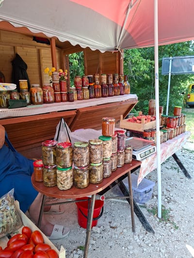 The image depicts an outdoor market stall in Scoreni, Moldova, on a sunny day. The stall is set up under a red and grey awning, supported by a white pole. In the foreground, a person wearing a blue garment and grey sandals is seated on the left. In front of them is a wooden crate filled with ripe red tomatoes. To the right, a red bucket is visible.
The stall itself features several tables laden with jars of preserved food. The top shelf displays jars of pickles, jams, and other preserved fruits and vegetables. A bouquet of yellow flowers is also placed on this shelf. The lower tables are stacked with more jars, predominantly containing preserved mushrooms and what appear to be pickled vegetables. A digital scale is placed on one of the lower tables. A wooden crate filled with peaches is also visible on the table.
In the background, there is a wooden market structure and lush green trees. A sign is partially visible in the distance, and a yellow vehicle can be seen parked further away. The ground is covered with gravel and small stones. The overall scene suggests a vibrant local market offering homemade preserved goods.