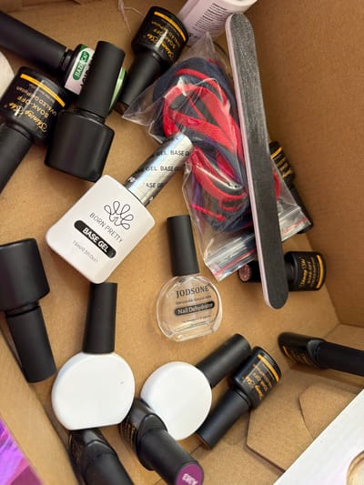 The image depicts a cardboard box filled with various nail care items. These include numerous bottles of gel nail polish in black bottles, some labeled "Shining She SOAK-OFF UV&LED Gel Polish", "JIAZHICAI", and a white bottle labeled "BORN PRETTY BASE GEL". A clear bottle of "JODSONE Nail Dehydrator" is also visible. Additionally, there's a black nail file, and a plastic bag containing blue and red laces. The specific location in Bearpark, United Kingdom, cannot be determined from the image.