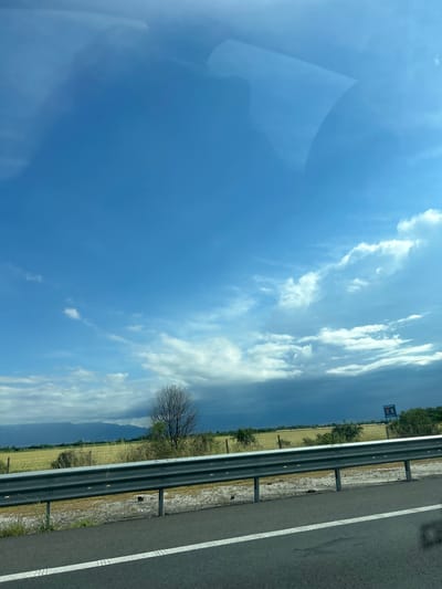 The image displays a sunlit rural landscape observed from a moving vehicle, with the photograph itself oriented vertically (rotated 90 degrees clockwise from its natural landscape perspective). The scene is dominated by a vast blue sky, featuring numerous scattered white cumulus clouds that indicate fair weather, likely during late morning or early afternoon.
On the right side of the image, a paved highway or road surface is visible, marked with a solid white line and a broken white line. A dark metal guardrail runs parallel to the road, separating it from a strip of dry grass and dirt shoulder. Beyond the guardrail, verdant fields stretch into the middle distance, intermingled with some patches of golden vegetation. Several trees are scattered across the landscape, including a prominent bare-branched tree positioned near the guardrail. In the far distance, faint outlines of mountains or hills are visible under a slightly hazy sky.
A blue rectangular sign with white text is discernible in the distance, partly obscured by foliage. The letters "BA" or "BД" appear at the top of the sign, suggesting it is a Bulgarian road sign or advertisement. The top-left corner of the image, when viewed in its current rotation, shows a reflection or glare from a window, consistent with the perspective from inside a vehicle. The overall scene conveys a sense of travel through an open, agricultural area near Malo Konare, Bulgaria.