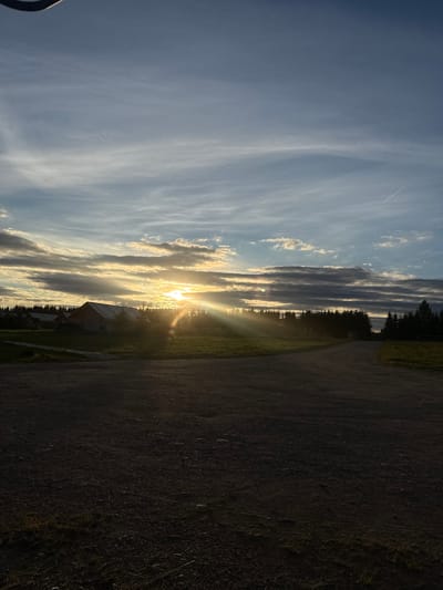 The image depicts a rural landscape at sunset in Zamosh'e (Zaklinskoe S/P), Russia. The foreground is a dark, gravelly dirt road. Beyond the road, a grassy field stretches towards a line of dark evergreen trees. In the middle ground, a cluster of buildings, possibly houses or cabins, are silhouetted against the sky. The sun is low on the horizon, partially obscured by dramatic, layered clouds, casting long rays of golden light across the scene. The sky is a mix of blues and grays, with wispy clouds and a visible contrail from an aircraft. The overall impression is one of a peaceful, natural setting at dusk.