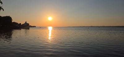 This is a panoramic, high-angle, wide shot of a serene lake at sunset, with the sun positioned slightly left of center, casting a brilliant orange glow across the water and sky. The scene evokes a sense of tranquility and natural beauty.

The foreground is dominated by the calm lake surface, reflecting the warm hues of the sunset with subtle ripples creating a textured pattern. Scattered across the water are a few buoys and possibly some floating debris.

In the middle ground, on the left side of the frame, a distinctive modern building with a triangular, metallic roof structure stands on the shore, suggesting a developed waterfront area. Adjacent to this building, trees and some smaller structures line the bank, creating a silhouetted effect against the bright sky. A pier extends from the shore into the water.

The background features a distant, low-lying shoreline, possibly a continuation of the land or a mountain range, softened by the atmospheric haze of sunset. Several small sailboats and buoys are visible on the water in the distance, hinting at recreational activities.

The time of day is clearly sunset, indicated by the low sun and the warm, golden-orange light bathing the entire scene. The sky transitions from a bright orange near the horizon to a softer, pale blue towards the top of the frame, suggesting clear weather. The overall mood is peaceful and picturesque. There is no visible text in the image. The location context provided is Bregenz, Austria, which aligns with the waterfront architecture and lake setting.