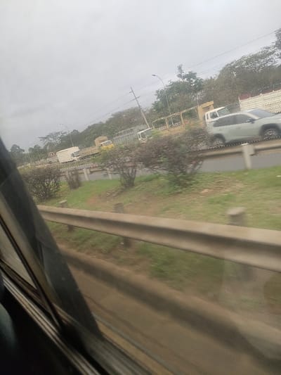 The image displays an exterior roadside view, captured from the interior of a moving vehicle, located in Kahawa Wendani ward, Kenya. A light-colored metal guardrail spans the lower portion of the frame, partially obscured by the vehicle's window frame on the left. Beyond the guardrail, a grassy median strip is visible, interspersed with several dark-foliaged bushes. A multi-lane highway occupies the midground, featuring vehicular traffic in motion. A light-grey SUV, possibly a Toyota RAV4, is prominent in the right lane, exhibiting motion blur. To the left and further down the road, a white cab-over-engine truck with a flatbed and a white box truck are observed, along with a yellow truck in the distant left lane. Utility poles supporting overhead wires and streetlights line the road parallel to the direction of travel. A dense treeline forms the background against an overcast, light-grey sky. No individuals are discernible in the scene.