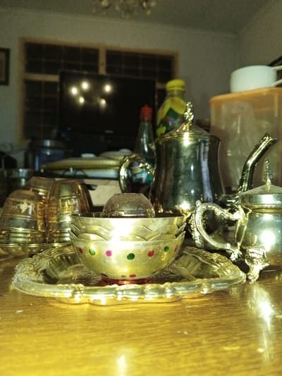 The image depicts an interior scene, likely a domestic setting, in Longmiang, Laos, focused on a collection of metalware on a polished wooden surface. In the immediate foreground, a round, ornate, gold-colored metal tray holds several items. A stack of small, decorative brass bowls is centered on the tray, with the topmost bowl featuring green and red polka dots on its exterior. To the left, several inverted brass cups are stacked. To the right, elements of an ornate, gold-colored metal teapot or creamer, including a handle and lid finial, are visible. Behind these items, a larger, tarnished silver-colored metal jug or teapot with a prominent lid and finial stands. Adjacent to it are a dark bottle with a red cap, a yellow-capped plastic bottle, and a light-brown rectangular plastic storage container with a white lid. In the background, a large, dark-screen television reflects several bright light sources. Behind the television, a window with a grid pattern is visible, framed by light-colored wood. A faint chandelier is discernible at the top center. No individuals are present in the scene.