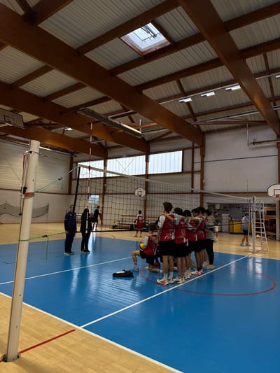 The image depicts an indoor sports gymnasium in Sennecey-le-Grand, France. A central volleyball court with a blue playing surface is visible, marked with white lines, while surrounding areas feature a wooden floor with red lines.
On the near side of the volleyball net, approximately seven to eight young males, primarily wearing red and white jerseys with numbers and black shorts, are gathered in a huddle, with arms around each other's shoulders. One individual in this group is kneeling. A pair of black shoes rests on the blue court near the kneeling individual.
To the left of the huddle, near the volleyball net's pole, an adult male in a blue long-sleeved shirt and dark pants stands, observing the group. Next to him, an adult female in dark clothing also stands.
In the background, behind the net, multiple individuals are present. One young male in a red jersey is visible further back on the court, near a basketball hoop. Another individual in a light blue shirt stands near a white equipment ladder or rack on the far right. Additional figures are partially visible further in the background.
The gymnasium features a high ceiling constructed with prominent wooden support beams and a corrugated metal roof. A square skylight is present in the ceiling, providing natural light. Fluorescent light fixtures are suspended from the ceiling beams. Large rectangular windows with multiple panes run along the upper sections of the walls, allowing additional natural light. Basketball hoops with backboards are mounted on the walls at both ends of the court. The volleyball net spans the width of the court, held by white poles secured into the floor.