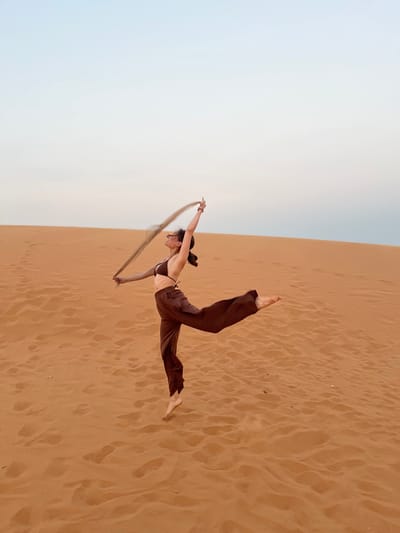 A solo female figure is depicted in mid-air, performing a dance or jump on an expansive reddish-orange sand dune, characteristic of the Mui Ne sand dunes near Phan Thiet, Vietnam. The individual is positioned left of center, viewed in profile facing left with her head tilted upward and a subtle smile. She wears a dark brown bikini top and matching loose, wide-leg trousers. Her dark hair is gathered behind her head. Her left arm is raised and bent, holding a light, flowing, grayish fabric or scarf that extends upward and backward. Her right arm is extended upwards and backward. Her left leg is bent at the knee with the foot positioned behind, while her right leg is extended backward, parallel to the ground, with the foot pointed. Both feet are bare. The sand surface exhibits ripples and scattered footprints. The background consists of further sand dunes receding into the distance, meeting a pale, light blue to off-white sky.