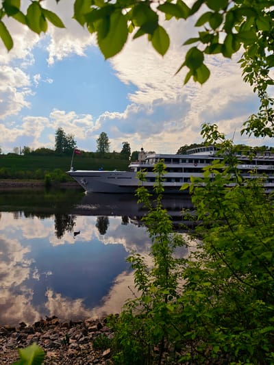 A bright, sunlit day in Водник Снт, Russia, features a large white passenger cruise ship sailing on a calm river. The ship, partially obscured by foliage in the foreground, is reflected in the river's surface, which mirrors the blue sky filled with puffy white clouds. Green trees line the riverbank, and a few buildings are visible in the distance. The foreground shows rocky shoreline partially covered by lush green undergrowth. Leaves from overhanging trees frame the top of the image.