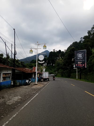 The image depicts a rural mountainous road in Ciloto, Indonesia, under an overcast sky. The asphalt road, marked with a solid white line on the left shoulder and broken yellow double lines in the center, recedes into the distance. On the left side, a dirt and gravel shoulder borders a series of single-story commercial structures with weathered corrugated metal roofs. The leftmost building features a blue lower facade, a white upper section, and a display window. A black and white scooter is parked on the dirt in front of it.
Further along the left shoulder, a decorative white street lamp post with gold finials and three ornamental lantern-like lights stands prominently. A circular white sign with Arabic script and "AL KUNA" in Latin letters hangs from this pole. Behind the pole, a white box truck is parked.
On the road, a dark-colored motorcycle or scooter with a single rider is traveling away from the camera in the right lane. On the right side of the road, a large black billboard displaying "ROOF PARK" in white and red text, with an arrow indicating "4.7 KM," is mounted on a dark pole. The background is dominated by a densely forested green mountain range under the cloudy sky. Overhead utility lines are visible crossing the upper portion of the frame from left to right.