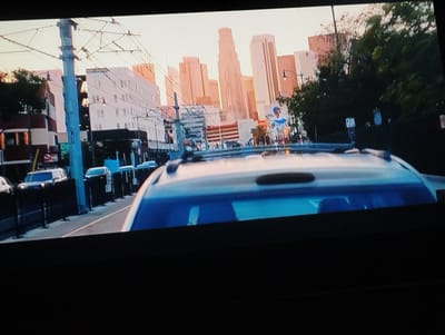 The image shows a view from inside a car, looking out toward a cityscape. The car's roof is visible in the foreground. In the mid-ground, there is a street with parked cars and sidewalks lined with black fences and utility poles with overhead wires. The background features a skyline of tall buildings, possibly located in Los Angeles based on the architecture. There is also a billboard visible in the mid-ground.