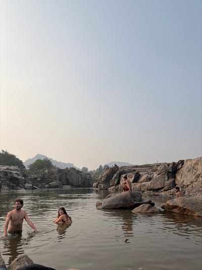 The image portrays a natural river environment in Hale Bandi Harlapura, India, under a hazy sky that transitions from a pale yellow-white near the horizon to a faint blue overhead. The scene focuses on a calm, brownish-green river bordered by extensive formations of large, grey, weathered rocks.
Four individuals are present in the foreground and midground. In the left foreground, a shirtless man with dark hair and a beard stands waist-deep in the water, facing slightly right. To his immediate right, a woman with long dark hair is partially submerged in the water, wearing a dark-colored top, and facing left. In the midground, a shirtless man wearing red shorts crouches on a large, partially submerged rock, looking down towards the water. Further to the right, a fourth shirtless man with dark hair and a beard is seated on a rock near the water's edge, partially obscured. Several small, dark figures are visible atop the rock formations along the right bank. The left bank consists of dense rock formations, with distant green foliage and hills visible beneath the hazy sky.