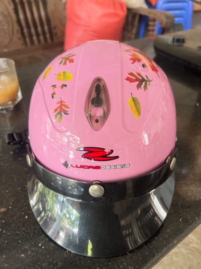 A pink motorcycle helmet with autumn leaf decals and a "LUCAS DESIGNS" logo sits on a dark, speckled countertop. A glass containing an amber liquid with ice is to the left of the helmet. In the blurred background, there is a red bag, a blue chair, and other indistinct objects. The image appears to be taken indoors, likely in a residential setting in Vị Xuyên Commune, Vietnam, though no specific landmarks confirm this location.