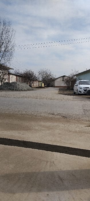 An eye-level, angled shot captures a road scene. A light-colored vehicle sits parked on the left side of the frame. The road itself occupies much of the view, with patches of gravel visible along its edges. Buildings line the left side of the road, partially obscured by bare trees. The sky above is overcast, filled with light blue and gray clouds, marked with what appears to be contrails. A concrete structure with two lines or shadows sits in the right side of the image.