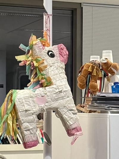 The image, oriented vertically, depicts an indoor scene featuring a unicorn piñata and a stuffed animal.

The primary subject is a white unicorn piñata, meticulously crafted from crinkled white tissue paper. It features a pink mane and horn, along with a vibrant, multicolored tail and a ruffled collar made of light pink, blue, yellow, green, and orange tissue paper. A black and white eye sticker is visible on its head, and a small pink heart adorns its side. The piñata is suspended by a string from a white horizontal bar or shelf on the left side of the frame.

Positioned above and slightly behind the piñata is a brown stuffed animal, resembling a camel or horse. It is adorned with a green, red, and white striped scarf and a small cap that clearly displays the text "DUBAI." This stuffed animal rests on a white shelf or within a storage unit located in the upper right portion of the image.

The background includes a dark, rectangular area on the left, likely a window or glass door, showing faint vertical lines. Above this, a light-colored wall or blinds with horizontal slats is visible. To the right, behind the stuffed animal, white shelving units hold various items, including a blue container. The scene suggests an indoor setting, possibly a home or office, indicating a playful or celebratory context. The time of day appears to be daytime, though no direct light source is prominent. There are no people visible. The given location context of Issy-les-Moulineaux, France, is not visually identifiable within the image.