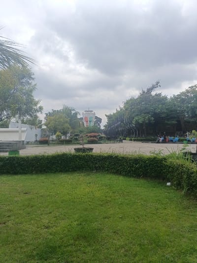 The image depicts a public park or plaza in thành phố Biên Hòa, Vietnam, under an overcast sky. In the foreground, a manicured lawn of green grass occupies the lower half of the frame, bordered by a low, dense, dark green hedge running horizontally across the mid-ground. Beyond the hedge lies a large, grey paved area, forming a central plaza.
To the far left of the plaza, a white concrete structure with visible steps is present. Scattered across the plaza are several circular planters containing small bushes, and a larger, darker planter holding a small, bare-branched tree. In the center-right, an abstract, dark-colored sculptural element stands, composed of multiple curved, vertical fins. Further right, along the edge of the paved area, several colorful, low-profile benches or seating units are visible in shades of blue, orange, and red. Approximately 3-5 indistinct human figures are seated or standing near these colorful units, appearing to be gathered.
The background is dominated by lush green trees with dense foliage, framing the plaza. A prominent, multi-colored water tower is visible in the distant center-left, featuring a white base and top, with vertical bands of green, orange, and possibly other hues in its cylindrical mid-section. The sky above is entirely covered with thick, grey clouds, indicating an overcast condition.