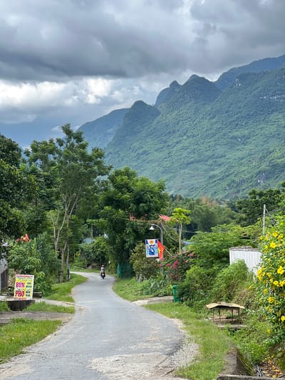 The image depicts a rural road in Lùng Tám Commune, Vietnam, under a cloudy sky. The road curves to the right, leading towards a lush green mountain range covered in dense vegetation. On the left side of the road, a sign for "Ngan Ha Noodle Soup Bun Pho" is visible. Further down the road, a person is riding a scooter, heading away from the camera. Various trees and foliage line both sides of the road, with a cluster of bright yellow flowers blooming on the right. A small, makeshift table with a thatched roof sits on the right side of the road, near a drain. The scene conveys a sense of remote, natural beauty, typical of the Vietnamese countryside.