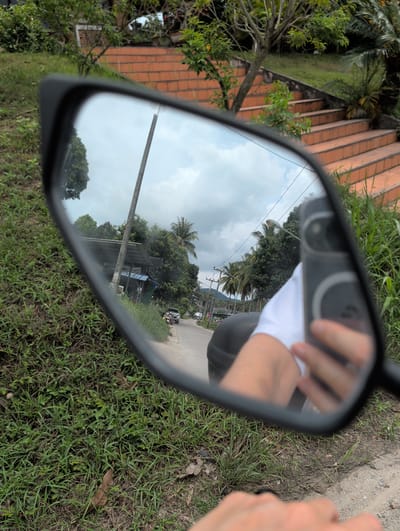 The image shows a motorcycle mirror reflecting a road in Moo 4, Thailand. The reflection includes a view of the road, trees, utility poles, buildings, and the sky. The person taking the picture and holding their phone is also visible in the mirror. The foreground surrounding the mirror consists of grass and dirt.