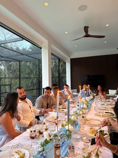 The image depicts an indoor dining event, likely a dinner party or celebration, set in a modern and elegant space. Several adults, both men and women, are gathered around a long dining table, conversing and enjoying a meal.
The table is elaborately set with a white tablecloth, a light blue fabric runner, and individual place settings. Each setting features ornate gold-colored charger plates, white main plates with food (including dishes with vegetables and what appears to be pasta or a casserole), ribbed clear glasses, and gold cutlery. Tall candlesticks with lit candles are spaced along the table, accompanied by decorative floral arrangements featuring white and light blue flowers. Small bottles, possibly of rosé wine, are placed at intervals, with some displaying indistinct text on their labels.
The dining area is characterized by a high ceiling with multiple recessed lights and a large, dark wooden three-bladed ceiling fan. Expansive floor-to-ceiling windows line one side of the room, offering a view of lush outdoor greenery, including palm trees, suggesting a warm climate. A dark, textured wall panel is visible in the background, contrasting with the lighter interior elements. The lighting indicates it's likely late afternoon or early evening, with natural light filtering through the windows complemented by artificial lighting and the warm glow of candles. The attendees appear relaxed and engaged, contributing to a pleasant social atmosphere. The modern architecture and tropical foliage are consistent with a setting in Naples, United States.