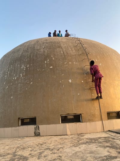 An outdoor scene in Barako, Nigeria, features a large, weathered, light-beige concrete dome structure under a clear, pale blue sky. A fixed metal ladder, comprising vertical rails and horizontal rungs, is attached to the dome's exterior, extending from the lower section towards its apex. One individual, dressed in a red and white patterned short-sleeved top and matching trousers, and wearing dark sandals, is actively ascending this ladder, positioned on its lower-mid section. Five other individuals, appearing to be adult males in various traditional garments including tunics and caps, are gathered at the dome's summit, appearing to sit or stand. The lower portion of the dome is encircled by a band of light-colored tiles, some exhibiting damage or misalignment, above which are several rectangular, unglazed openings. The foreground presents a flat, textured concrete ground surface. The lighting indicates clear ambient daylight, with warmer illumination evident on the right side of the dome.
