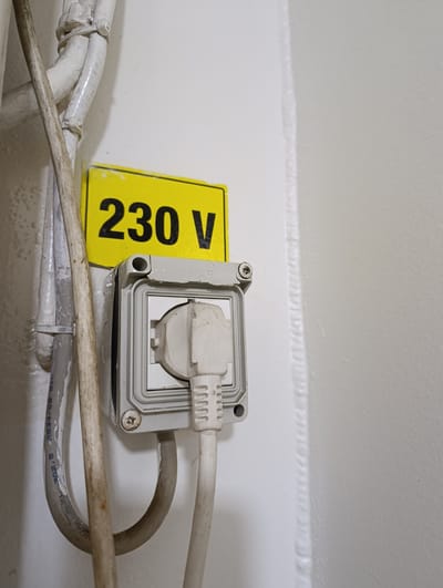 A close-up, eye-level shot depicts a wall-mounted electrical outlet and a yellow warning sign in Šabac, Serbia. The outlet is covered by a gray, rugged housing with a clear plastic lid. A white electrical plug is inserted into the outlet. To the left of the outlet, a yellow rectangular sign with black text reads "230 V", indicating the voltage. Several white and gray electrical cables are visible, running vertically along the wall and connected to the outlet housing. The wall appears to be painted a light color, possibly white or off-white, and shows some texture. The lighting is neutral, with no strong shadows. The scene is indoors, likely in an industrial or utilitarian space, given the type of outlet and the warning sign.