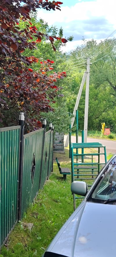 In Altayskoye, Russia, this outdoor scene features a green corrugated metal fence running along the left side of the frame, adorned with four black posts topped with white and black outdoor lights. The fence has diamond-shaped cutouts. To the right of the fence, a wooden park bench sits on the grass. Beyond the bench, a green metal playground structure with wooden seating and horizontal bars is visible. A white utility pole with power lines stands prominently in the background, amidst lush green trees. A portion of a silver car is in the lower right corner of the image, with its side mirror clearly visible. A paved road is discernible in the background, to the right of the utility pole, and a yellow sign with red text is partially visible on the right side of the road. The sky is partly cloudy with white clouds. The ground is covered in green grass and some patches of dirt.