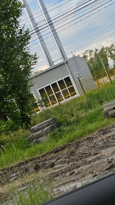 The image depicts an industrial area with a gray building, possibly a small factory or utility structure, with large windows. Two tall, metallic chimneys rise from the roof of the building. The building is surrounded by green grass, some of which is overgrown. In the foreground, there are stacks of concrete blocks, suggesting ongoing construction or renovation. A dirt road with tire tracks and some standing water is also visible in the foreground. Overhead, power lines crisscross the sky. In the background, there is a yellow fence and more trees. The image appears to be taken in an outdoor, semi-rural or industrial setting. The sky is cloudy. There are no people visible in the image. The location is identified as Kamensk-Uralsky, Russia.
