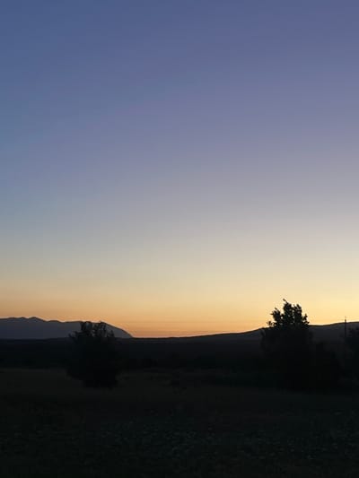 The image shows a dusk or dawn landscape, likely in Gorna Kremena, Bulgaria. The sky transitions from a pale blue to a gradient of orange and yellow near the horizon. A dark landmass with trees and possible vegetation is silhouetted against the bright sky. A thin, dark line likely represents a mountain range in the distance. No people, animals, or distinct objects are visible other than the natural elements.