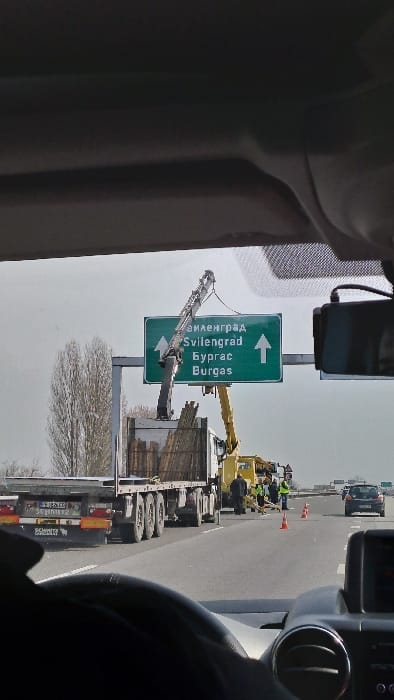 The scene captures a highway in Voysil, Bulgaria, during daytime under an overcast sky, viewed from within a vehicle. An overhead green highway sign displays "Svilengrad" and "Burgas" in both Cyrillic and Latin script, accompanied by upward arrows. On the right shoulder or partially in the rightmost lane, a white semi-truck with a flatbed trailer is stationary, loaded with numerous bundles of wooden poles. A yellow truck-mounted crane is positioned adjacent to the semi-truck, its boom extended over the cargo, indicating active loading or unloading. Three individuals, attired in high-visibility yellow/lime-green vests, are present near the crane and truck. Orange traffic cones delineate the work area on the asphalt pavement, with one cone horizontally positioned. A black passenger vehicle is observed in the distance in the leftmost lane. Deciduous trees with bare branches are visible to the left of the road. The immediate foreground includes portions of a vehicle dashboard, steering wheel, and a windshield-mounted device.