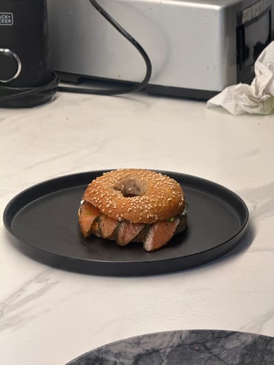 A bagel with salmon is presented on a matte black plate. The plate is set on a white marble countertop. A Black+Decker appliance is partially visible on the left. A crumpled white paper towel lies near the top left corner. The location is likely a kitchen in Levallois-Perret, France.