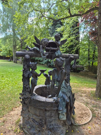 This is a medium shot of a bronze sculpture depicting a well with cherubs and a woman, located in Reisbach, Germany. The sculpture is weathered and has a greenish patina. The well is made of stone and is surrounded by grass and fallen leaves. The woman is seated on the edge of the well, looking up. Cherubs are positioned around the well, one pouring water from a shell and others interacting with a bucket and pulley system. Trees with green and some red foliage form the backdrop, with a wooden fence visible in the distance. The overall setting appears to be a park or garden.