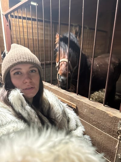The image depicts an indoor scene within a stable located in Kalinovo, Russia. In the foreground, a female individual is captured in a selfie-style photograph. She wears a light brown knit beanie and a voluminous, light-colored fur or faux fur garment. Her long, dark hair is visible beneath the beanie. Behind a barrier of vertical metal bars, a brown horse with a dark mane and a light-colored halter is visible inside a stall. The stall's walls are constructed from wooden planks, and bedding material is present on the floor. Artificial lighting illuminates the upper portion of the scene. The female's gaze is directed towards the camera, while the horse's head is slightly lowered, facing generally forward.