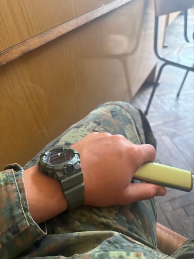 A man wearing a military-style camouflage uniform and a green G-Shock watch is sitting at a wooden table. He is holding a small, rectangular yellow device in his hand. The table and a chair are visible in the background. I have no information about the location of Dolna Mitropolia, Bulgaria.