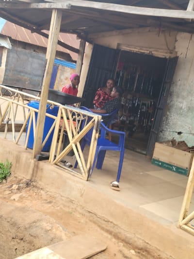 In the city of Bokkos, Nigeria, a low-angle, medium shot captures a scene outside a small shop. The shop's entrance is a dark opening with metal bars, revealing shelves stocked with items inside. In front of the shop, a blue plastic chair is occupied by a person wearing a black and white patterned shirt, facing left. To their right, another person in a red and black patterned shirt is seated, looking towards the first person. A third individual, wearing a red shirt and a pink headscarf, stands behind a black tray, looking towards the viewer. Yellow metal railings run along the foreground, with a large blue plastic bin positioned behind them to the left. The ground is a mixture of dirt and concrete. A cardboard box with "X Preston" written on it rests on the ground to the right. The roof of the shop is constructed of wood and corrugated metal.