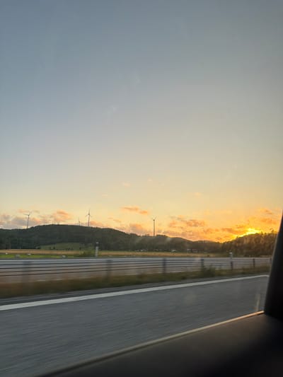 The image shows a highway landscape at sunset with wind turbines on a hill in the background. The sky is a gradient of blue and orange, with some scattered clouds. The hill is covered with trees and some fields. There are several wind turbines visible on the top of the hill. The highway is in the foreground, with lanes marked by white lines. The overall impression is one of a serene and scenic rural landscape. The specific location in Dingle, Sweden is not identifiable from the image alone.